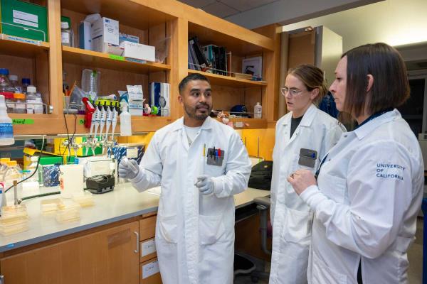 Chris Bravo, Margot Bacino, and Sue Lynch wear white lab coats as they discuss their work at the Sue Lynch lab.